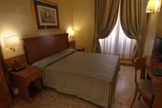 A double bed with a quilted grey cover sits between two wooden bedside tables, with a desk and television on the right and a curtained window behind.