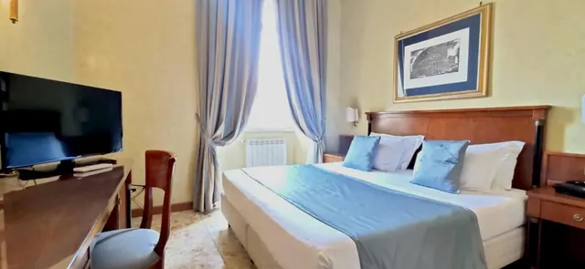 A hotel double bedroom with a blue-covered bed, a wall-mounted flat-screen TV, a wooden desk and chair, and a framed print of the Colosseum on the wall.