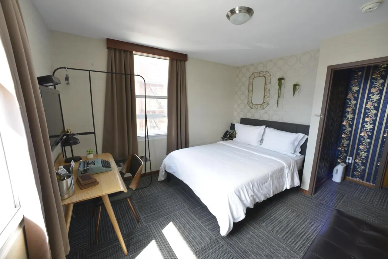 A bedroom with a king bed, a wooden desk with a typewriter and chair, a garment rack, and a window facing a brick building.