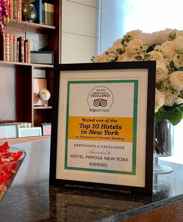 A framed TripAdvisor Certificate of Excellence stands on a reception desk beside a bowl of candy and a tall vase of white flowers.