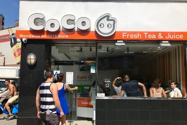 Street-level storefront of a CoCo Fresh Tea & Juice shop, with people standing at the entrance and customers ordering inside the glass front.