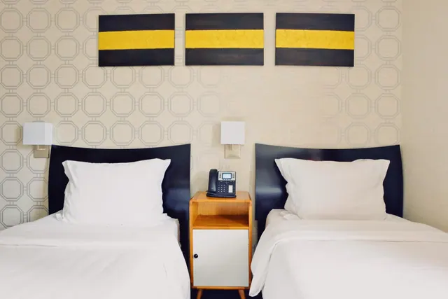 Two single beds with white bedding sit against a patterned wall, with a bedside table holding a telephone between them and three striped paintings hang above.