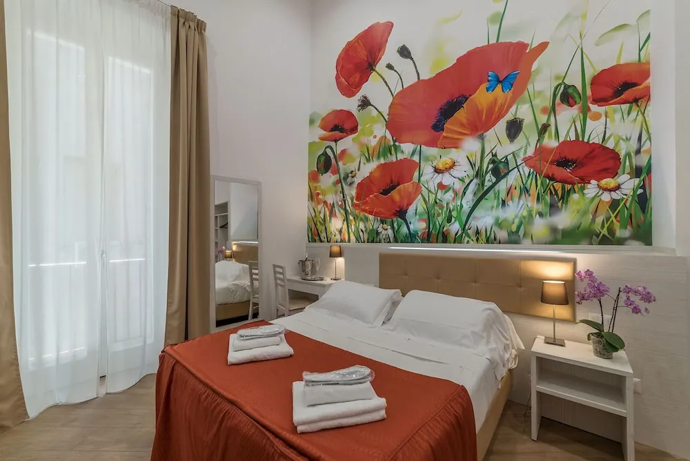 A double bed with a red throw and folded towels stands in a room with a large poppy mural on the wall, a nightstand with an orchid, and sheer curtains over the window.