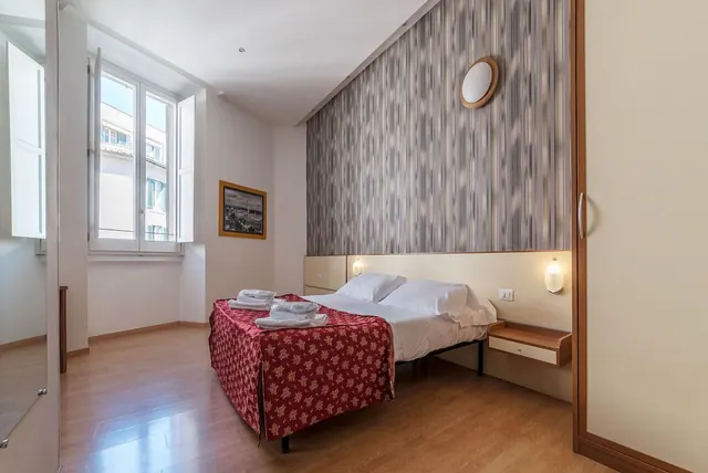 A bedroom with a double bed under a red patterned throw with folded towels, a striped feature wall, and a window with white shutters.