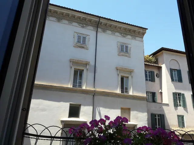 View through an open window of a cream-coloured building facade with green shutters, with purple flowers on a wrought iron railing in the foreground.