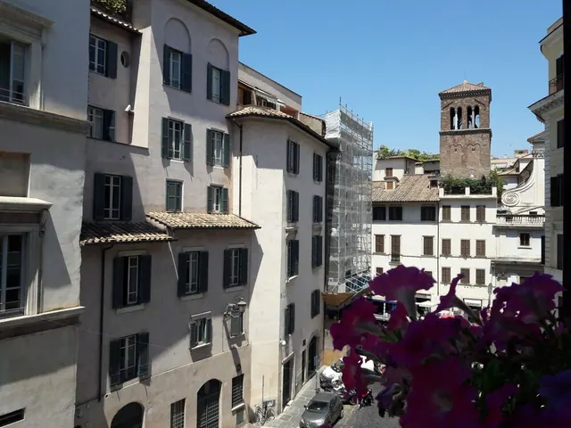 View from an upper floor down a narrow Roman street lined with old ochre and cream buildings, parked scooters and cars, scaffolding on one building, and a brick bell tower in the distance, with pink flowers in the foreground.