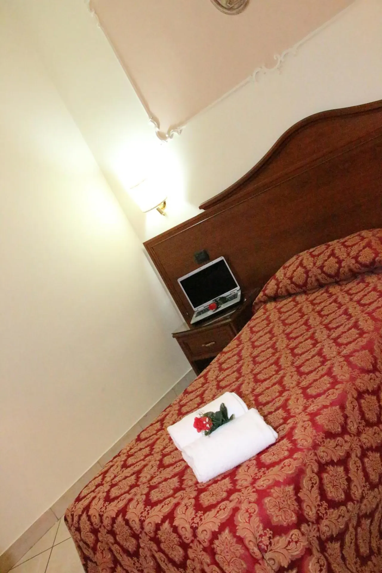 A double bed with a red patterned bedspread holds folded white towels and a red flower, with a small television on a wooden nightstand beside it and a wall lamp above.