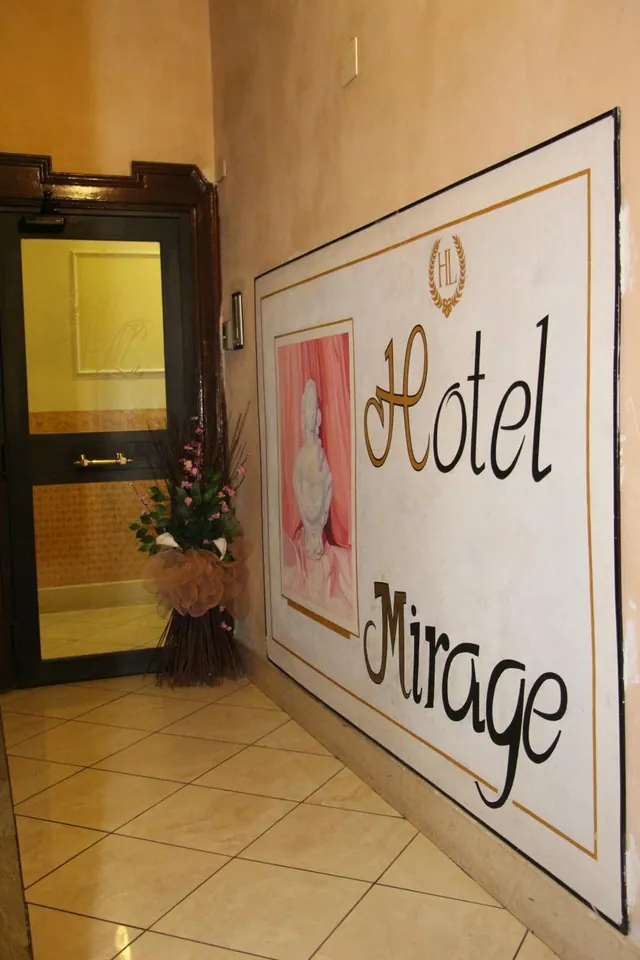 A hotel entrance hallway with a large painted wall sign reading Hotel Mirage, a glass door, a floral arrangement and a tiled floor.