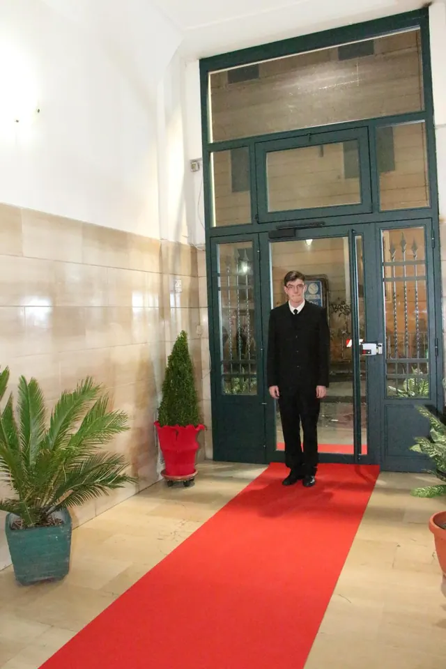 A man in a dark suit stands in a doorway of an entrance lobby with a red carpet runner and potted plants.