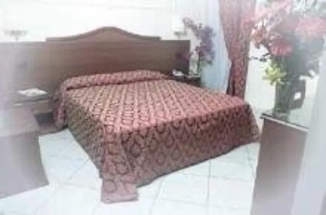 A double bed with a patterned red bedspread sits in front of a wooden headboard, with a white tiled floor and a vase of flowers on a side table beside it.