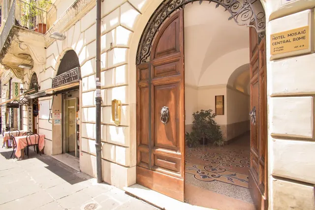 The entrance of Hotel Mosaic Central Rome, with open wooden doors, an ornate iron arch, a patterned tiled floor inside and outdoor tables with pink cloths on the street.