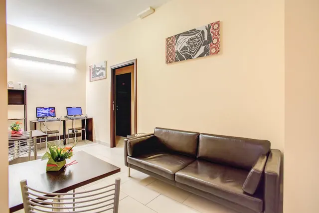 A living area with a brown leather sofa, a wooden dining table with metal chairs, and a desk with two computer monitors against a cream wall.