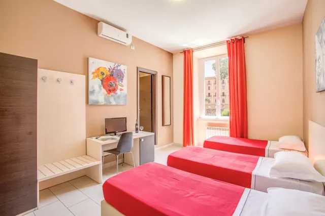 A bedroom with three single beds with red covers, a desk with a television and chair, a wall-mounted air conditioner, and a window with red curtains looking onto a building.