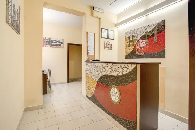 A reception area with a mosaic-covered front desk, a mosaic wall panel with the letters HM, cream walls and a tiled floor, with a doorway leading to a room with chairs behind.
