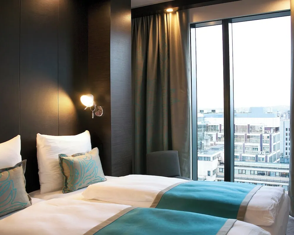 A bedroom with two single beds with teal runners, a wall lamp, and floor-to-ceiling windows looking out over city buildings.