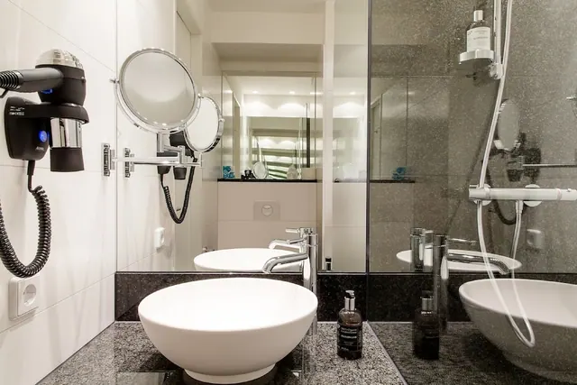 A modern bathroom with two round white basins, a glass-walled walk-in shower, a wall-mounted hairdryer and magnifying mirrors on a dark speckled stone surface.