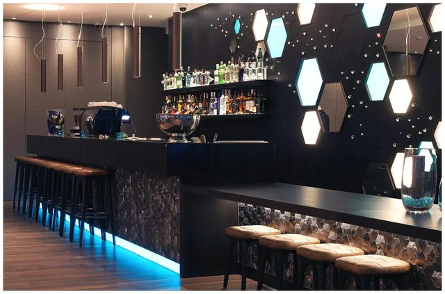 A bar counter with stools, shelves of bottles and illuminated hexagonal panels on a dark wall.