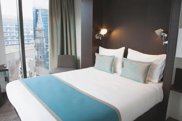 A made king bed with a teal runner and cushions sits beside floor-to-ceiling windows showing city buildings and a church tower.