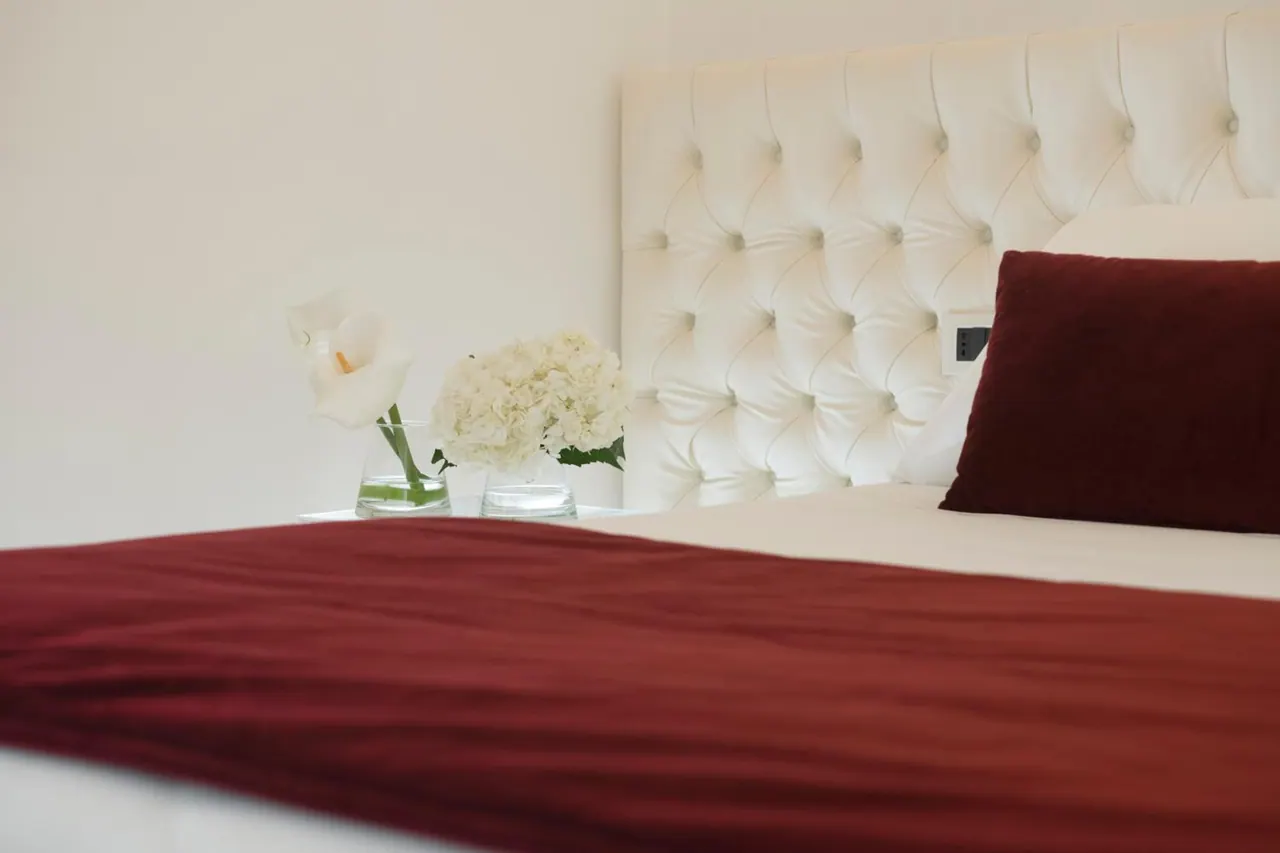 A made bed with a tufted white headboard, a red throw and cushion, and a nightstand holding white flowers in glass vases.