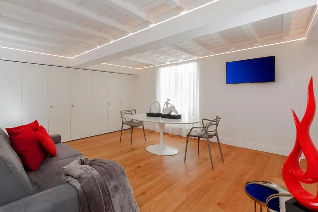 A living room with a grey sofa and red cushions, a white round table with two grey chairs, a wall-mounted television, white built-in wardrobes and a beamed ceiling with LED strip lighting.