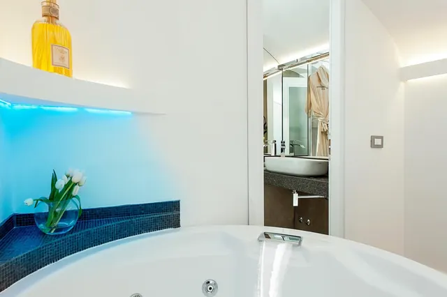 A white bathtub with jet fittings sits in the foreground beside a blue-lit tiled niche holding tulips, with a sink and hanging bathrobe visible through the doorway.