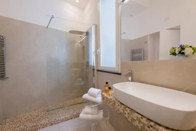 A bathroom with a glass-walled walk-in shower, an oval vessel sink on a mosaic-tiled counter, stacked towels and a frosted window.