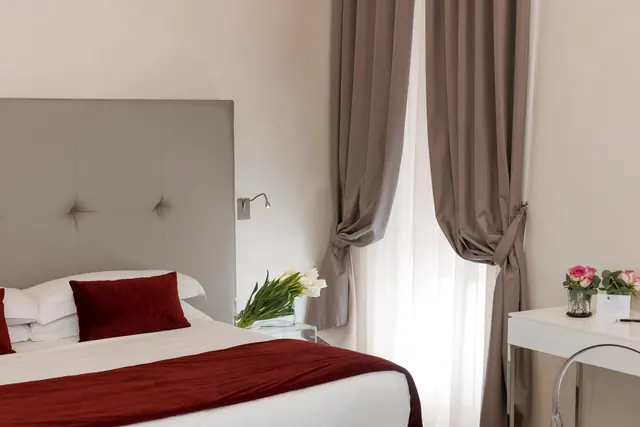 A made double bed with a grey tufted headboard and red throw sits beside a vase of white tulips, with a white desk holding pink roses on the right.