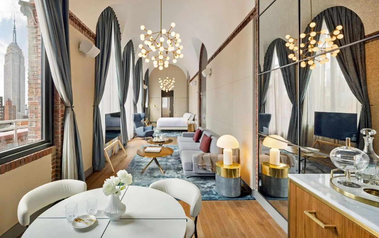 A bright suite living area with a grey sofa, round coffee table, tall arched windows, and the Empire State Building visible outside.