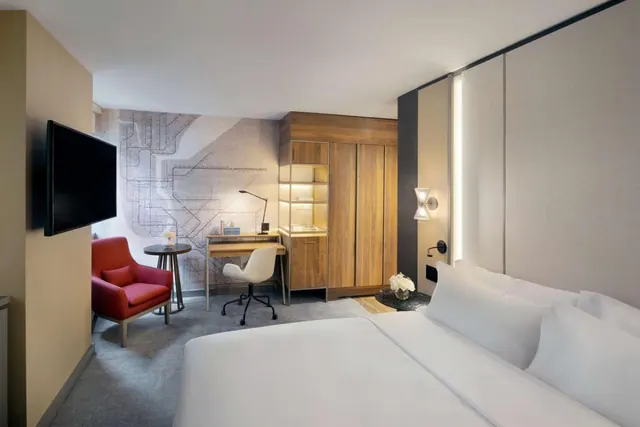 A hotel bedroom with a large white bed, a wooden desk with a white chair, a red armchair, a wall-mounted television and a wood wardrobe unit.