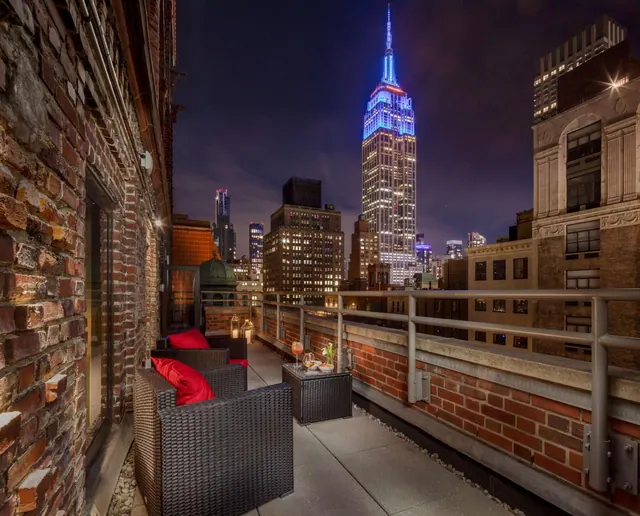 A rooftop terrace at night with wicker armchairs, red cushions and a side table, with the illuminated Empire State Building visible beyond the railing.