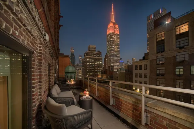 A rooftop terrace at dusk with outdoor lounge chairs, side tables and a railing, with the illuminated Empire State Building visible in the distance.
