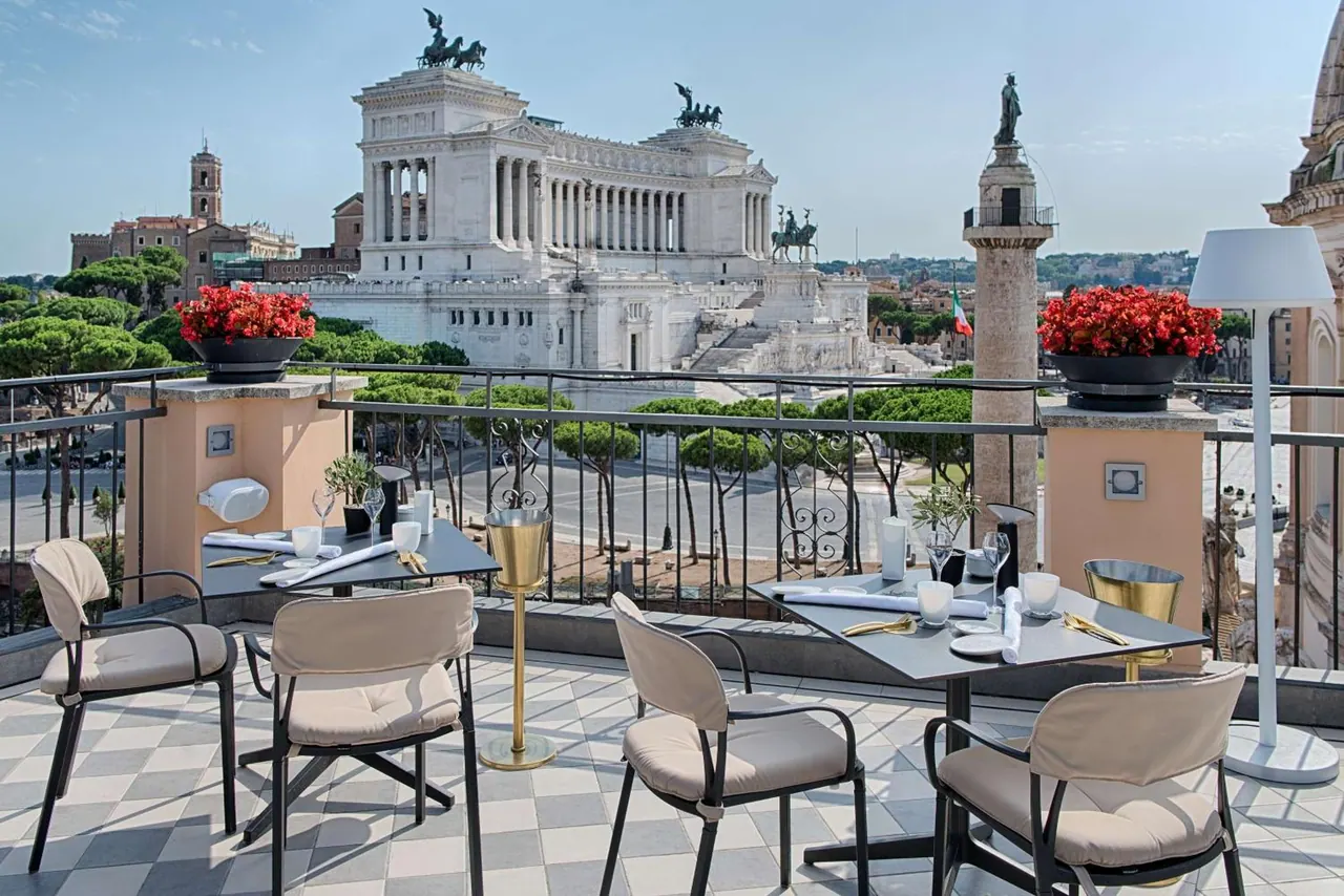 A rooftop terrace with set tables and chairs overlooks the white Vittoriano monument and Trajan's Column.