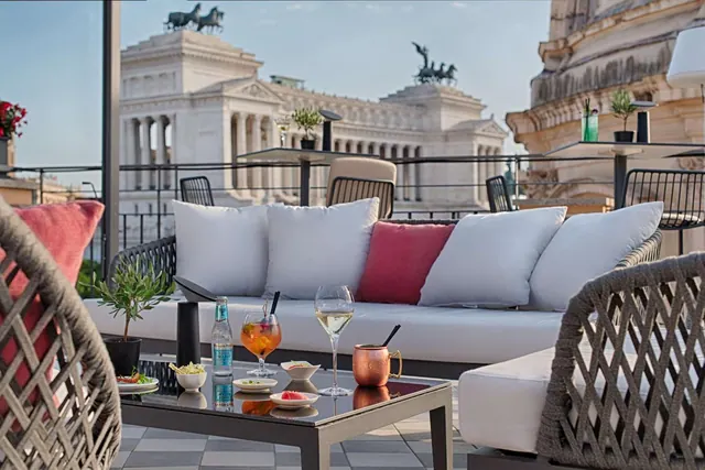 A rooftop terrace seating area with a white sofa, cushions and a glass coffee table holding drinks and small dishes, with the Vittoriano monument visible behind.