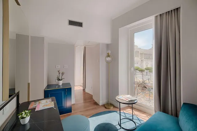A living area with a teal sofa and a round side table, and a tall window with curtains looking out toward the Vittoriano monument.