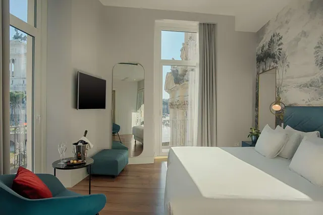 A bedroom with a large white bed, teal armchair and wall-mounted television, with tall windows looking onto a white classical monument.