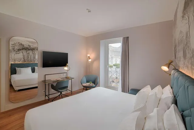 A hotel bedroom with a large white bed, a wall-mounted television, a desk and a blue armchair, with a window showing a view of the Vittoriano monument.