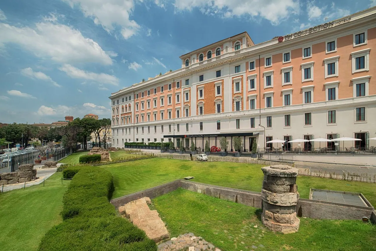 The exterior of a large cream and orange hotel building stands behind a lawn and ancient stone ruins under a partly cloudy sky.