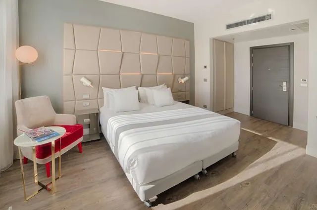 A bedroom with a large upholstered headboard, a made double bed, a red velvet armchair beside a small side table, a wardrobe and a closed door.