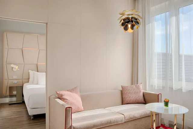 A beige sofa with pink patterned cushions sits beside a round side table and a sheer-curtained window, with a bed visible through the open doorway on the left.