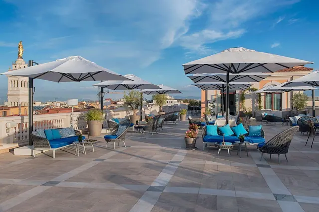 A rooftop terrace with grey parasol umbrellas, blue cushioned sofas and wicker armchairs, overlooking city rooftops with a golden statue on a tower to the left.