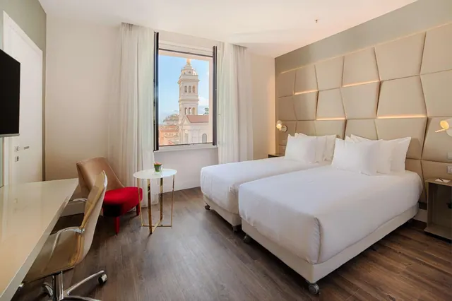 A hotel bedroom with two single beds, a desk, a red armchair and a window overlooking a church bell tower and rooftops.