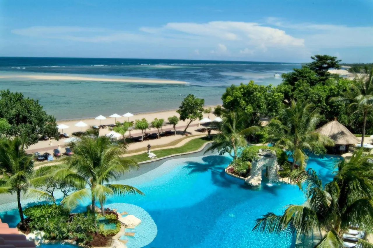 Elevated view of a large freeform swimming pool with a waterfall feature and palm trees, with a sandy beach, parasols and the sea beyond.