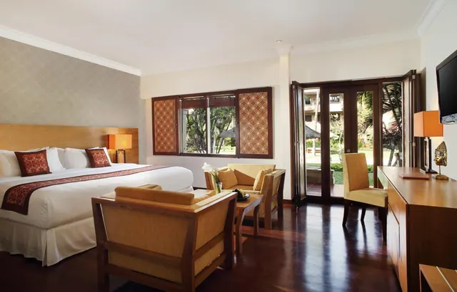 A bedroom with a king bed, an armchair and sofa seating area with a coffee table, a wall-mounted television, and glass doors opening onto a garden with palm trees.