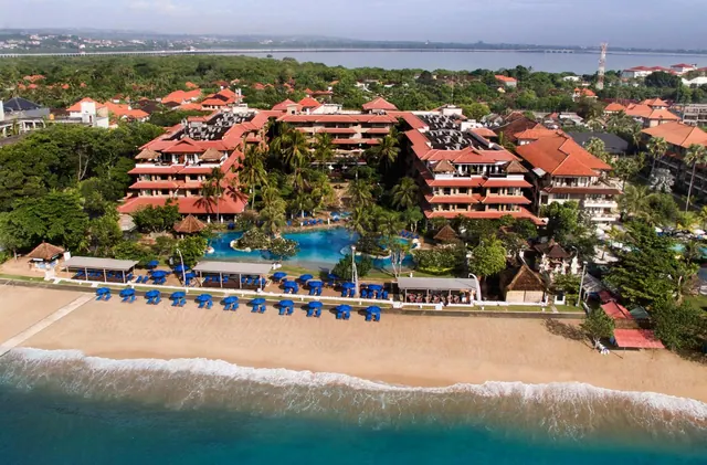Aerial view of a resort with terracotta-roofed buildings, a freeform pool, blue sun loungers and umbrellas along a sandy beach beside the sea.