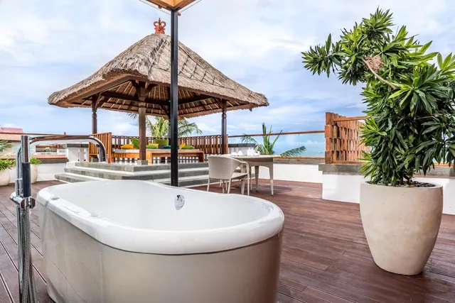 A freestanding bathtub sits on a timber rooftop terrace beside a thatched gazebo, a dining table with chairs, and the sea visible in the background.