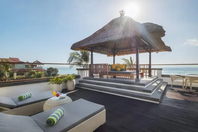 A rooftop terrace with a thatched gazebo containing a daybed, two grey sun loungers, a small table with drinks, and a view of the sea.