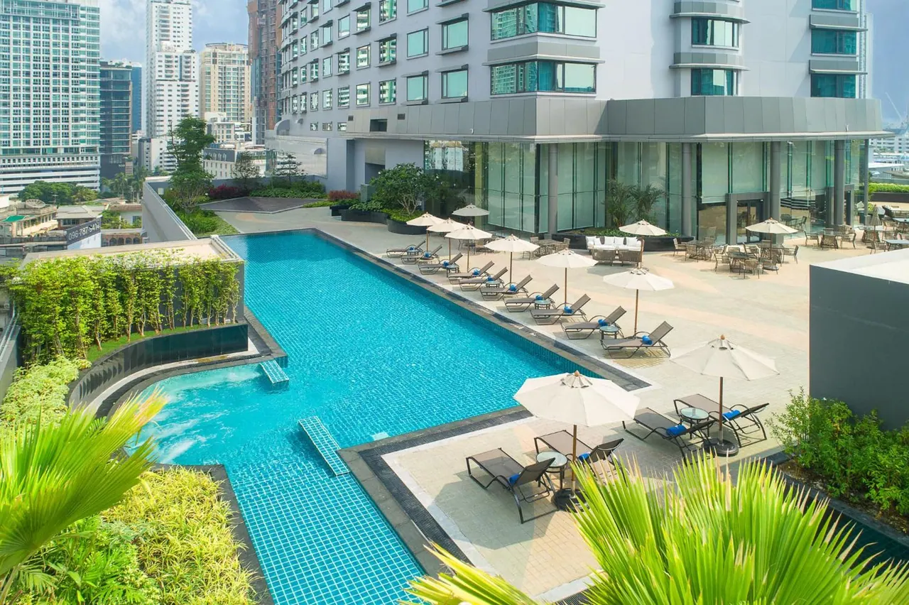 A long rectangular swimming pool with sun loungers and white parasols on a terrace beside a hotel building, with city towers in the background.
