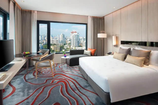A bedroom with a king bed, a desk with chairs, a grey sofa and a television, with a window showing a city skyline.