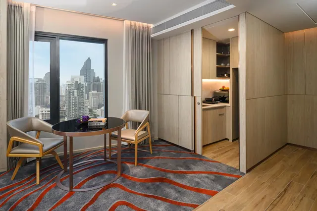 A hotel room with a round dark table and two upholstered chairs beside a window overlooking city skyscrapers, with a kitchenette visible beyond.