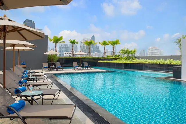 A rooftop swimming pool with sun loungers and shade umbrellas, with high-rise buildings and palm trees beyond.